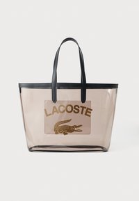 Prozirna torba s crnim ručkama, koja ima svijetlosmeđi obrub i smeđi Lacoste logo s grafikom krokodila sprijeda.
