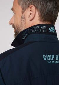 Camicia blu navy con colletto alto, caratterizzata da testo turchese che recita "Table Mountain P. peak 1.084,6 M" e dettaglio ricamato "Camp D.".