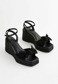 Sorte kile-sandaler med en platform-design, der har en vævet tekstur, to ankelstropper og en sløjfeaccent fortil.