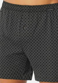 Zwarte zwemboxers met een textuurpatroon van witte geometrische vormen, elastische tailleband en een knoopdetail aan de voorkant. Lichtgewicht, sneldrogend materiaal.