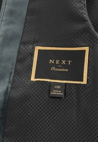 Schwarzes Stoffetikett mit goldener Schrift "NEXT Occasion" und Größe "40R"; strukturierter Hintergrund und zusätzlicher Tag mit der Angabe "Hergestellt in Bangladesch."