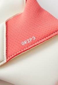 Korallszínű szövet, texturált mintával és fehér varrással, a "GRIP3" felirattal kiemelten megjelenítve. A háttér világos.
