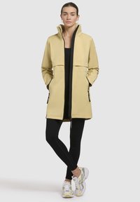 Ljusgul vattentålig trenchcoat med hög krage, svarta detaljer, två sidofickor och figurnära design som bärs över svarta leggings och sneakers.