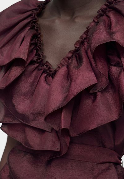 Κοντινό πλάνο ενός βαθύ βυσσινί φορέματος με υφή και μεγάλες βολάν γύρω από το V-λαιμόκοψη σε σκούρο δέρμα.