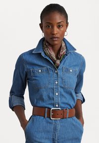Chemise en denim bleu clair avec un devant à boutons, deux poches poitrine et des manches retroussées, associée à une ceinture en cuir marron et un foulard à motifs.
