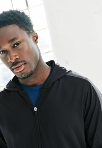 Musta vetoketjullinen huppari, jossa on sileä pinta ja huppu, pidettynä sinisen urheilupaidan päällä. Tyylikkäänä yksityiskohtana hopeanvärinen vetoketju ja hillityt yksityiskohdat olkapäillä.