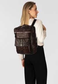 Zaino in pelle marrone con finitura strutturata, dotato di tasca frontale e dettagli con cerniere argentate, indossato su una spalla.