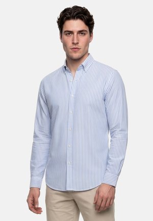 Homme portant une chemise à rayures verticales bleu clair et blanc avec boutons, et un pantalon beige, debout devant un fond uni.