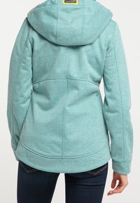 Chaqueta de forro polar en color verde azulado con capucha, que presenta un acabado texturizado, detalles de costura y un dobladillo ligeramente curvado. Combinada con vaqueros oscuros.