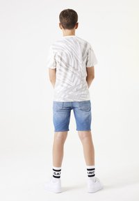Witte t-shirt met een subtiele bladerenpatroon, gecombineerd met lichtblauwe denimshorts, witte sneakers en zwartgestreepte sokken.