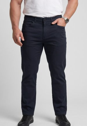 Man draagt donkerblauwe broek, wit t-shirt, zwarte sneakers, zilveren polshorloge, met één hand in de zak en de andere naast het lichaam.