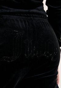 Närbild av svarta velourbyxor med glansiga svarta strassbokstäver som stavar "Juicy" på baksidan av höften.