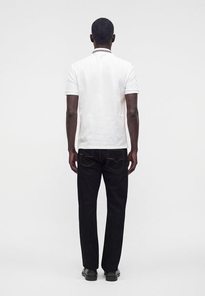 Homme debout, dos tourné, portant une chemise blanche à manches courtes, un jean noir avec des coutures orange et des chaussures noires sur un fond uni.