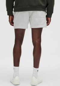 Shorts en coton gris au design simple, avec une poche arrière. Portés avec des chaussettes blanches et des baskets, sur un fond neutre.