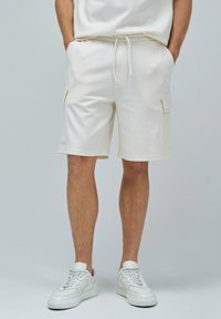 Shorts en coton blanc avec poches cargo sur les côtés, une taille à cordon de serrage et une coupe décontractée, associés à des baskets blanches.