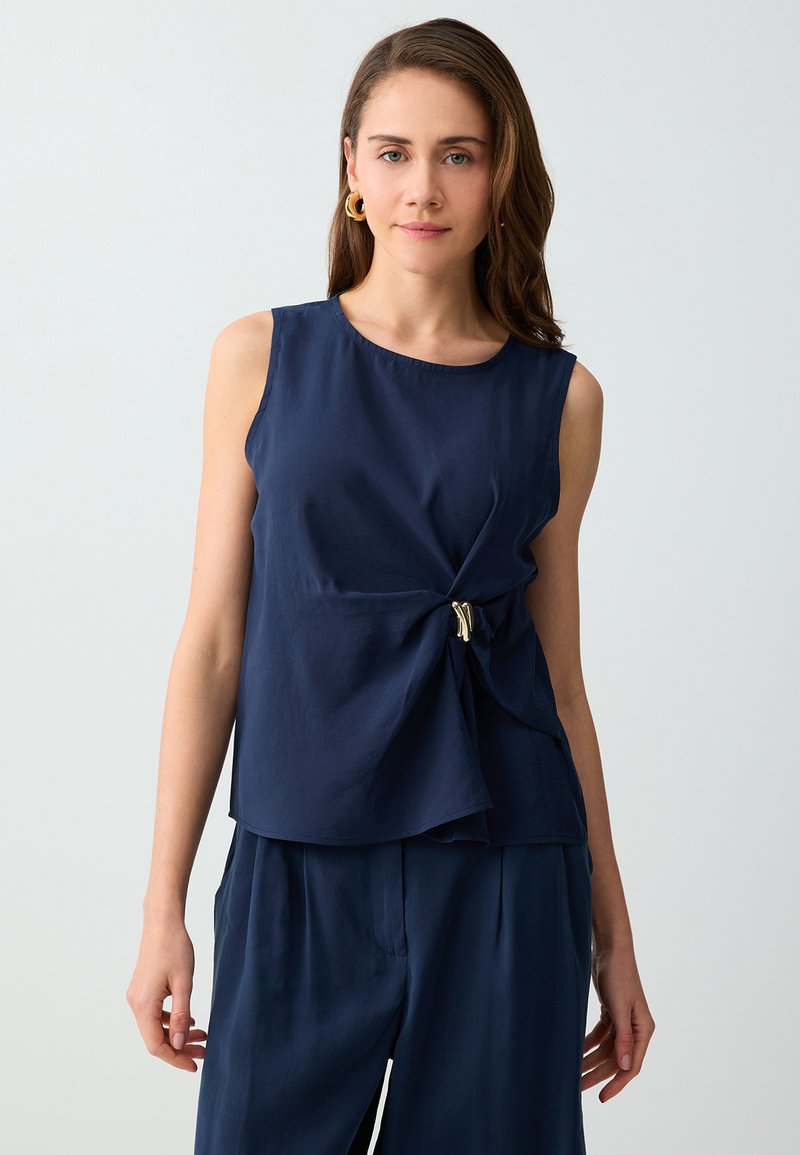 Femme aux cheveux bruns mi-longs portant des boucles d’oreilles anneaux dorées et un haut sans manches bleu marine avec une boucle latérale, assorti à un pantalon bleu marine.