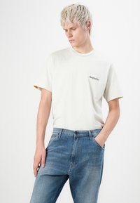 Ung mand med kort blond hår iført en hvid Replay Inc. T-shirt og blå jeans, med den ene hånd i lommen, kigger ned.