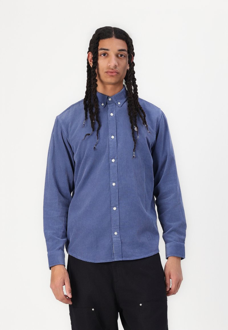Button-down shirt in blue fabric with a textured finish, featuring white buttons and long sleeves. Cutaway collar and rounded hem.