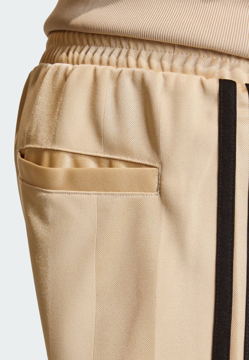 Pantalones deportivos beige con tela texturizada, que cuentan con una cintura elástica y un bolsillo lateral de diseño elegante, acentuados por rayas negras.
