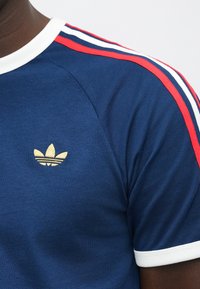 Artimos planas asmens, dėvinčio tamsiai mėlyną sportinį marškinėlį su baltomis ir raudonomis juostelėmis ant pečių ir mažuoju auksiniu Adidas logotipu ant krūtinės.
