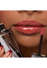 Lakier do ust Vinyl Lock w przezroczystej tubce, z czarną zakrętką. Błyszczące usta w delikatnym różowym odcieniu, z miękkim aplikatorem eksponującym produkt.