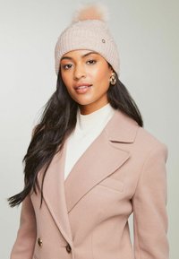 Căciulă tricotată roz deschis, cu textură în relief, având un pom-pom din blană artificială și accesorii din perlă. Pură cu o haină bej și un pulover cu guler înalt crem.