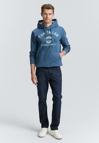 Hanorac albastru din bumbac cu imprimare cu logo, buzunar frontal și șnururi, asortat cu blugi albastru închis și adidași albi.