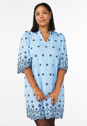 Naine kannab helesinist kleiti, millel on musta lilletikandit, seisab käed ees kokku pandud, valge taustaga.