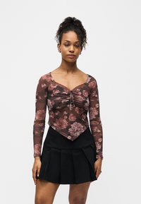 Νεαρή γυναίκα με σγουρά μαλλιά, ντυμένη με ροζ μπλούζα με μακριά μανίκια και φλοράλ σχέδιο και μαύρη πλισέ φούστα, που στέκεται μπροστά σε απλό λευκό φόντο.