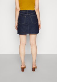 Saia de ganga azul escura com corte reto, bolsos traseiros visíveis e costura em contraste. Usada com uma blusa amarela e sandálias de salto castanhas.