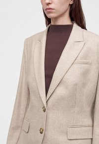 Femme portant un blazer beige par-dessus un haut marron côtelé à col montant, montrée de la poitrine au menton sur un fond blanc uni.