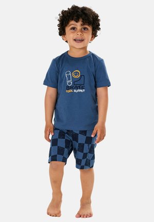 Toddler with curly hair wearing a blue t-shirt with skateboard print and blue checkered shorts, standing barefoot on a white background.
