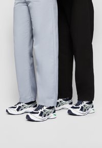 Två personer som står sida vid sida, klädda i vita, marinblå och gröna sneakers, där den ena har på sig ljusgrå byxor och den andra svarta byxor.