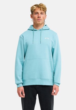 Giovane uomo con capelli biondi indossa una felpa con cappuccio azzurro chiaro con tasca frontale e pantaloni neri, in piedi davanti a uno sfondo bianco uniforme.