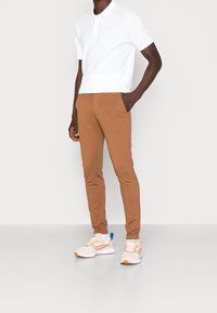 Polo blanc à manches courtes, pantalon slim marron, et baskets claires avec des accents orange. Tenue décontractée adaptée à diverses occasions.