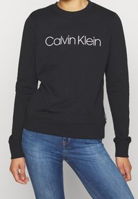 Felpa nera con colletto a girocollo, maniche lunghe e logo bianco "Calvin Klein" ben visibile sul davanti. Texture liscia.