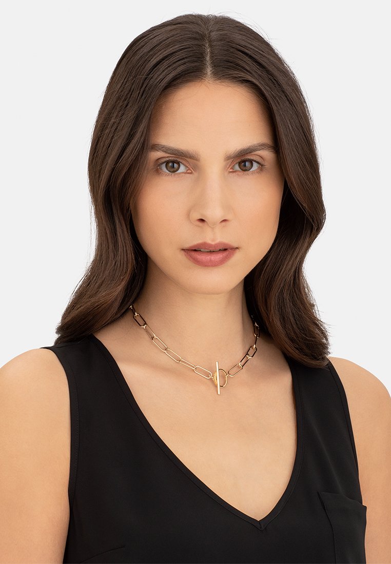 Guldtonet metalkæde med forlængede led og en toggle-lukning, hvilende om modellens hals, mod en ensfarvet baggrund.