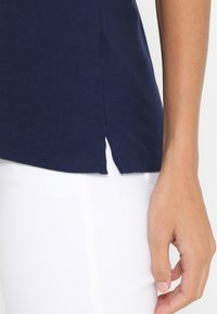 Marinblå bomullströja med sidoflik och rundad nederkant, kombinerad med vita byxor, som visar textur och tygdetaljer.