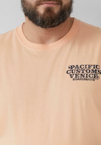 Hellbeige Baumwoll-T-Shirt mit Rundhalsausschnitt, mit dunkelblauer Textdruck "PACIFIC CUSTOMS VENICE BOARDS&KITS" auf der Brust.