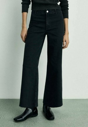 Personne portant un jean noir taille haute à jambes larges avec ourlets bruts et des bottines noires, debout devant un fond clair uni.