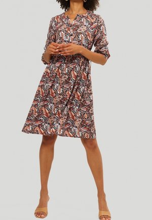Frau in knielangem Kleid mit mehrfarbigem Paisley-Muster und hochgekrempelten Ärmeln, steht mit locker verschränkten Händen.