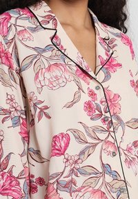 Top da pigiama floreale in morbido tessuto beige, con motivi di fiori rosa e viola, piping nero e dettagli con bottoni sul davanti.