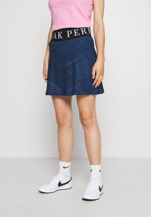 Donna con top rosa, gonna blu scuro con banda nera con scritte in vita, calzini bianchi Nike e sneakers bianche Nike, in piedi contro uno sfondo uniforme.