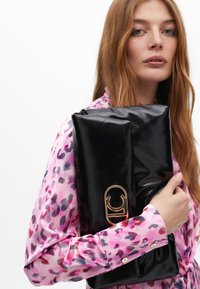 Model s dugom smeškom nosi sjajnu crnu torbu s zlatnom kopčom, obučena u vibrantnu ružičastu bluzu s šarenim uzorkom.