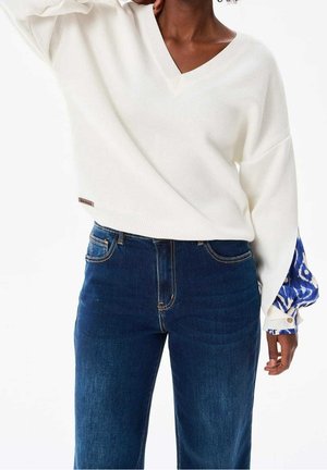 Person trägt locker sitzenden weißen V-Ausschnitt-Pullover mit blau gemusterten Ärmeln und hoch taillierte dunkelblaue Jeans, steht vor weißem Hintergrund.