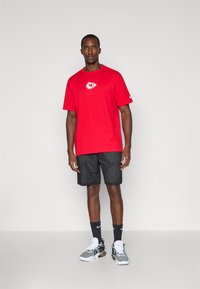 Sarkana T-krekls ar logo uz krūtīm, apvienots ar melniem sieta šortiem, melnām zeķēm un pelēkbaltiem sporta apaviem.