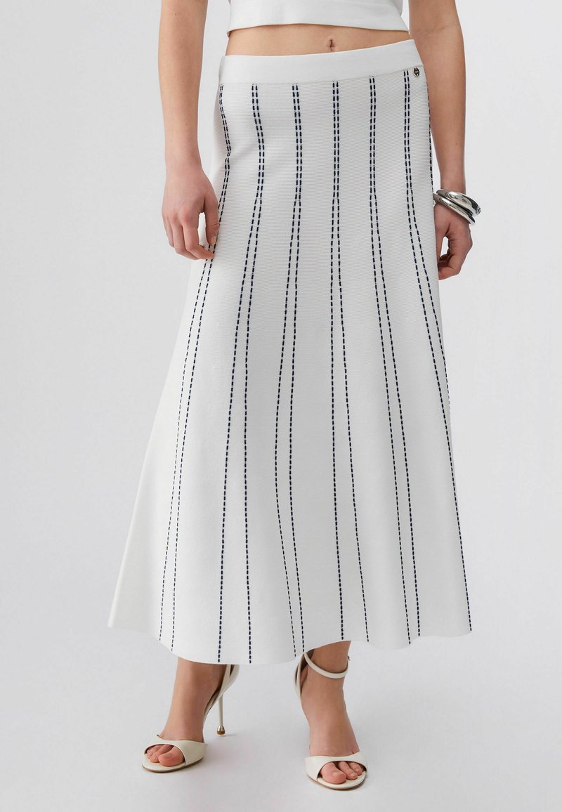 Valge A-lõikega seelik vertikaalsete mustade õmblusjoontega, kantud valgete avatud varbaga kontsade ja hõbedaste käevõrudega paremal randmel.