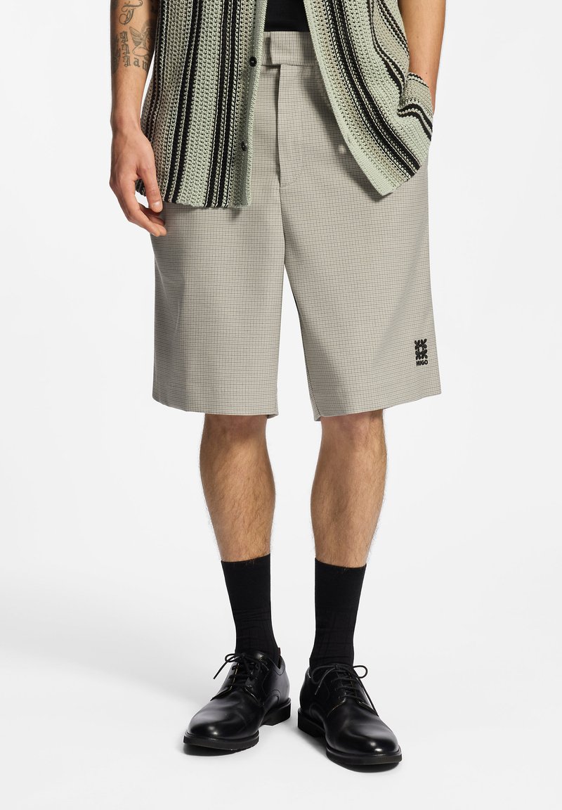 Homme portant un short beige à carreaux jusqu'aux genoux, des chaussettes noires, des chaussures habillées en cuir noir, et un gilet sans manches en maille avec des rayures noires et crème.