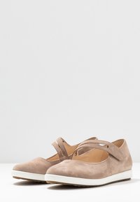 Zapatos Mary Jane de ante beige con una suela blanca plana, punta redondeada y detalle de tiras entrecruzadas en el empeine. Textura suave.