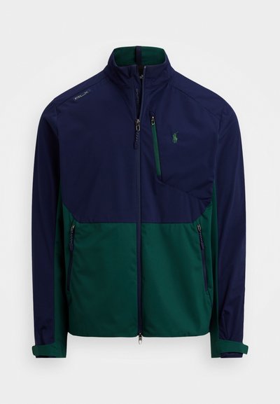 Ναυτική και πράσινη ζακέτα με φερμουάρ στις τσέπες, ψηλό γιακά και λογότυπο στο στήθος. Διαθέτει σχεδίαση με χρωματικές μπλοκ και μαλακό ύφασμα, καθώς και ρυθμιζόμενους καρπούς.